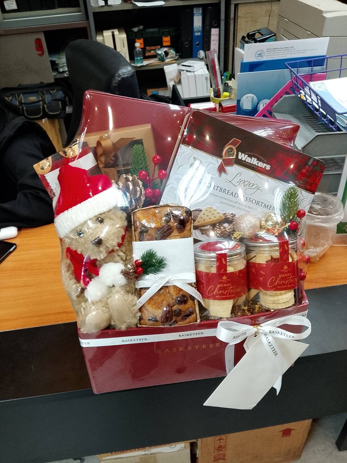A festive Christmas Cheer Gift Box sits on a desk, filled with a teddy bear in a Santa hat, Walkers shortbread, pastries, and two tins with Christmas designs, all adorned with pine sprigs and ribbons. Office items appear in the background.