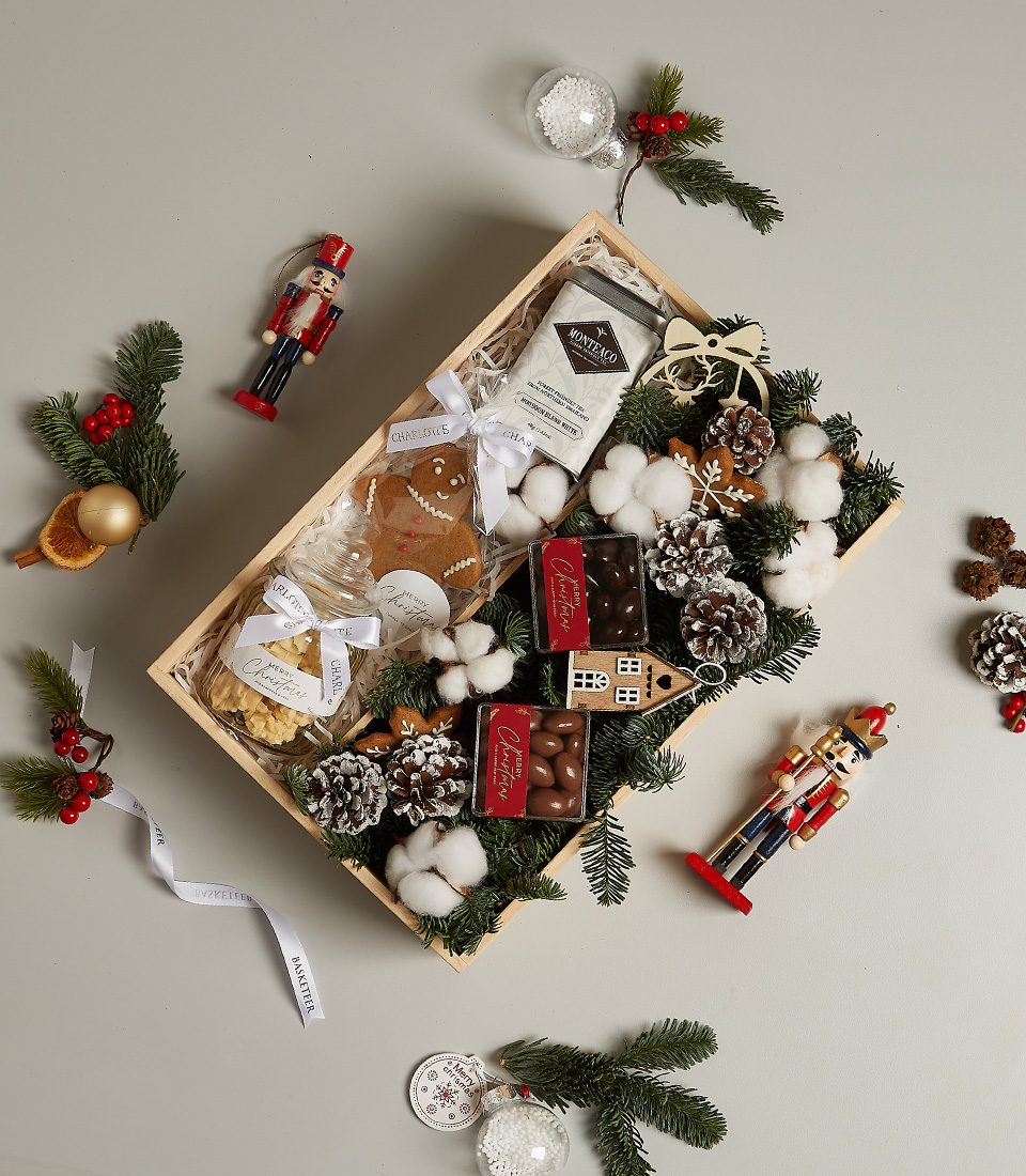 Festive gift box featuring gingerbread cookies, chocolates, hot cocoa, and holiday decor with pinecones and cotton snow.