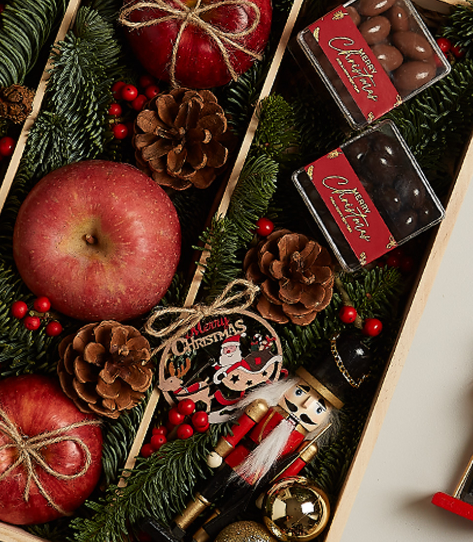 A Festive Cheer Deluxe Gift Box showcases a vibrant Christmas display with red apples, pinecones, evergreen branches, ornaments, and small boxes of chocolate. A wooden nutcracker, red berries, and a 