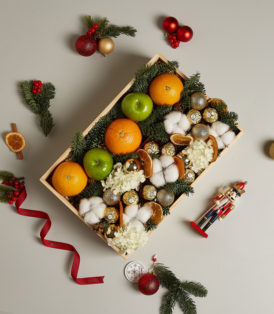 Holiday gift box with fresh oranges, green apples, Ferrero Rocher chocolates, dried oranges, and festive decorations.