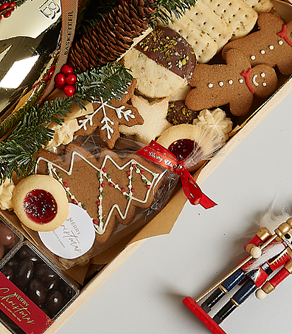 A festive arrangement of holiday cookies featuring gingerbread, pastries, and a tree-shaped cookie with icing. Garnished with pine, holly, and a nutcracker figure. A ribbon ties a bag of treats in this golden celebration cookie & champagne gift box delight.