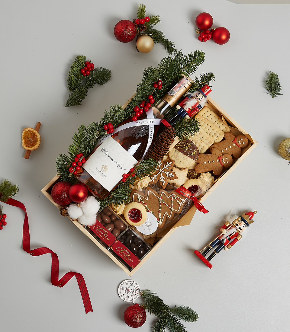 Festive gift box with rosé wine, holiday cookies, gingerbread, chocolates, seasonal greenery, and decorative nutcracker ornaments.
