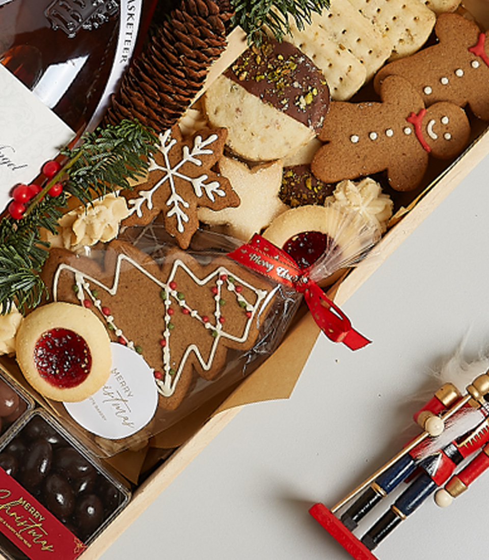 Festive gift box with rosé wine, holiday cookies, gingerbread, chocolates, seasonal greenery, and decorative nutcracker ornaments.