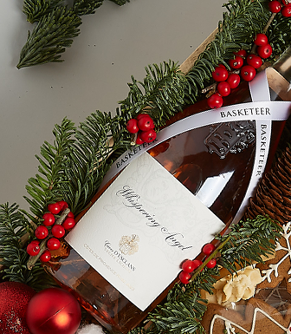 Festive gift box with rosé wine, holiday cookies, gingerbread, chocolates, seasonal greenery, and decorative nutcracker ornaments.