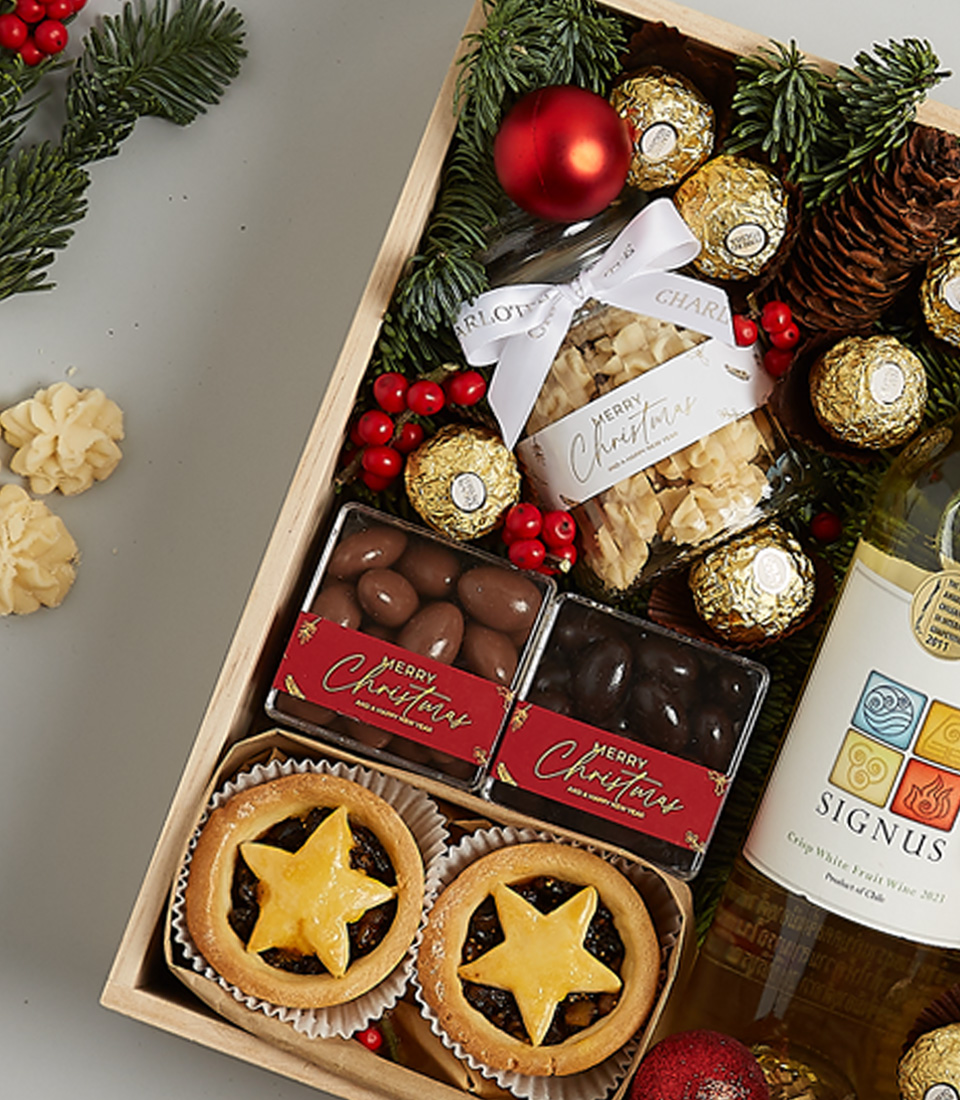 Christmas gift hamper featuring Signus white wine, Ferrero Rocher chocolates, mince pies, gingerbread cookies, and festive greenery with ornaments.