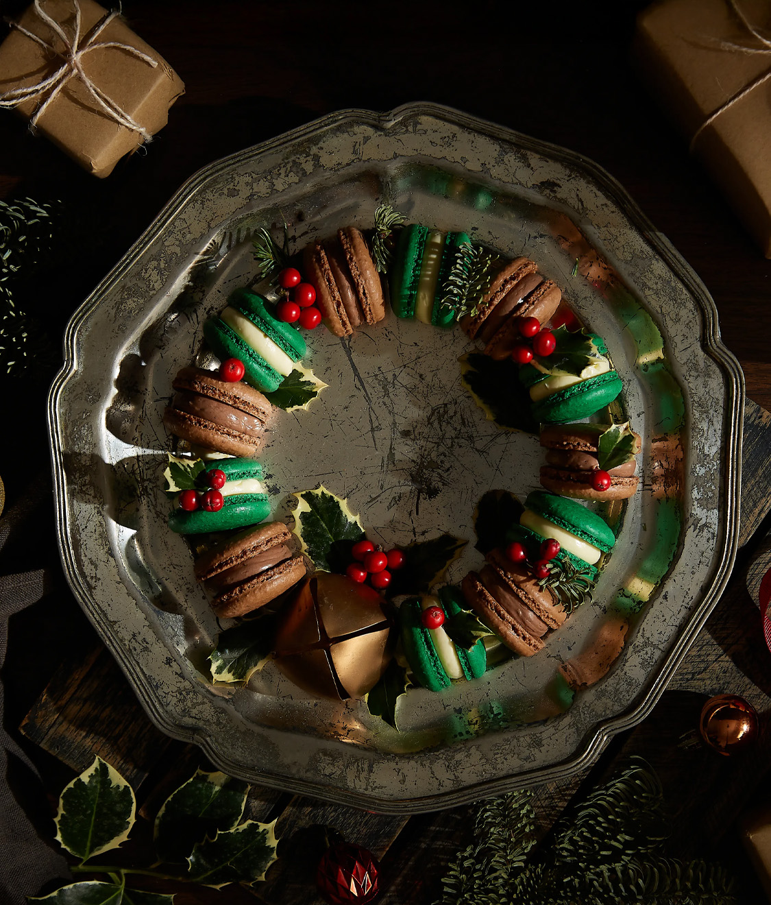 A decorative macaron wreath featuring chocolate and green macarons adorned with holly leaves and red berries, beautifully displayed on a rustic metal platter.