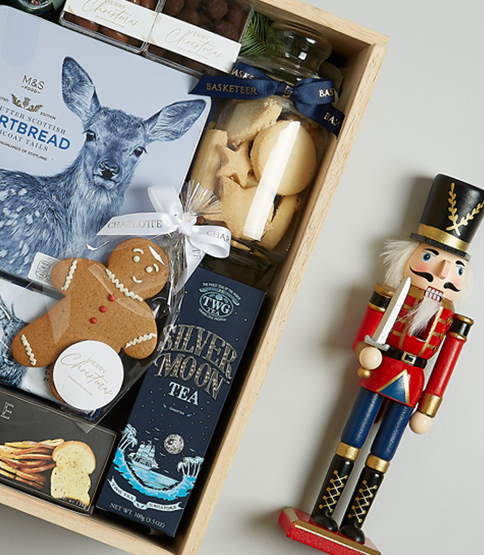 Luxurious Christmas gift basket with sparkling wine, shortbread, black truffle crisps, Silver Moon tea, chocolate almonds, and a gingerbread cookie in a festive blue theme.