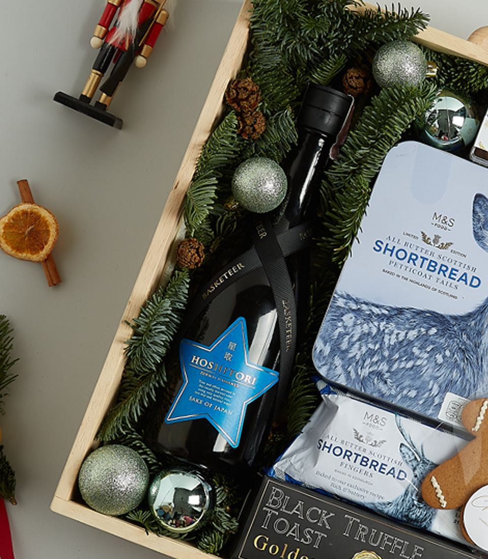 Luxurious Christmas gift basket with sparkling wine, shortbread, black truffle crisps, Silver Moon tea, chocolate almonds, and a gingerbread cookie in a festive blue theme.