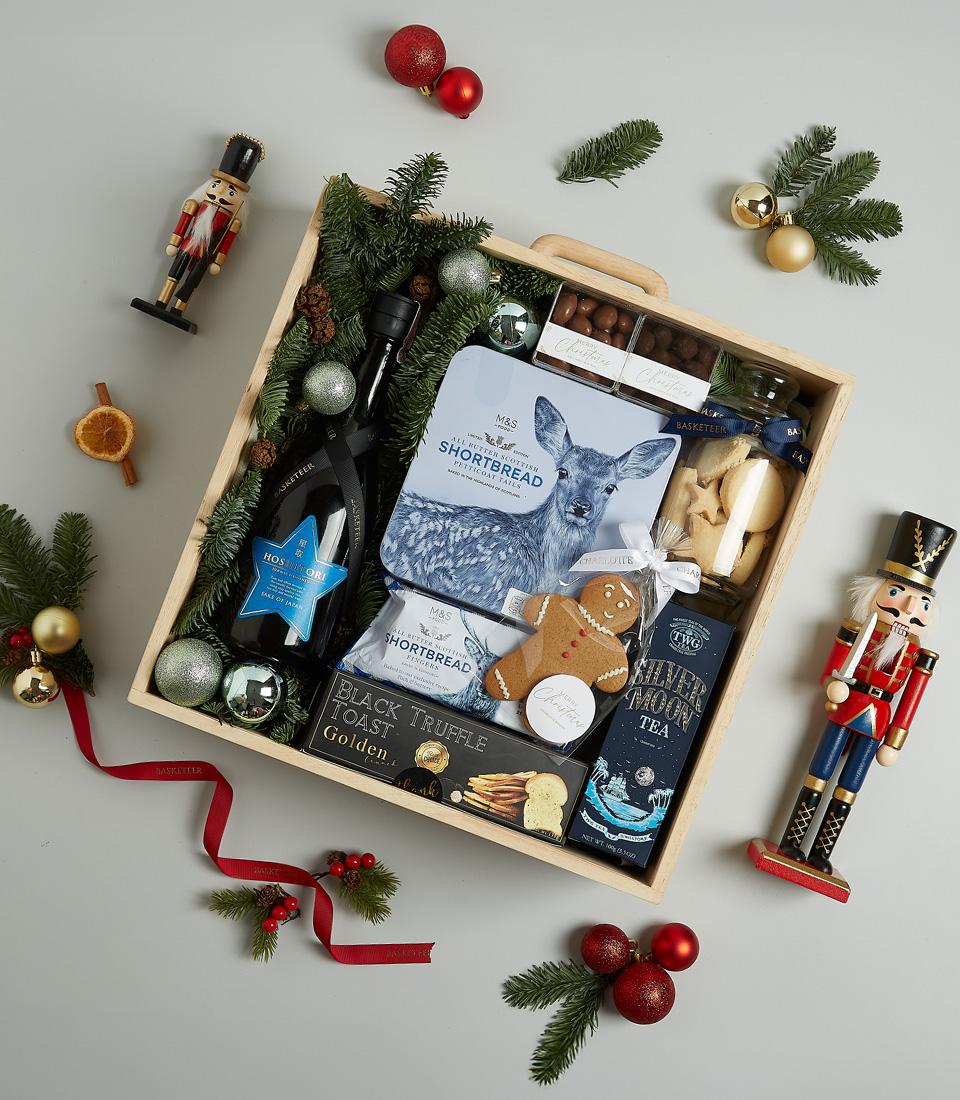 Luxurious Christmas gift basket with sparkling wine, shortbread, black truffle crisps, Silver Moon tea, chocolate almonds, and a gingerbread cookie in a festive blue theme.