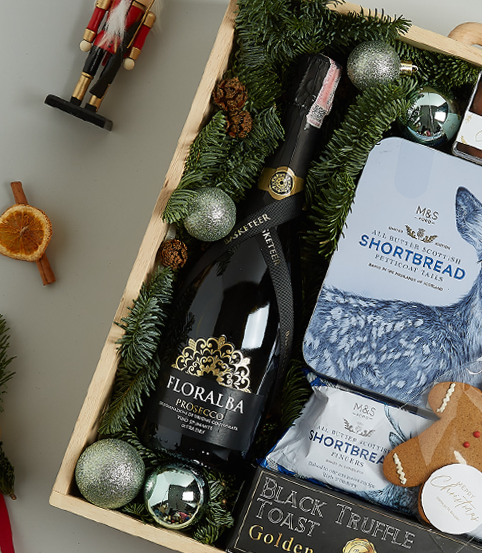Luxurious Christmas gift basket with Floralba sparkling wine, shortbread, black truffle crisps, Silver Moon tea, chocolate almonds, and a gingerbread cookie in a festive blue and green theme.