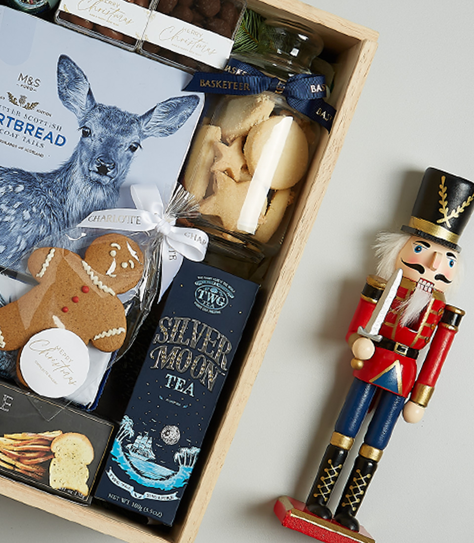 Luxurious Christmas gift basket with Floralba sparkling wine, shortbread, black truffle crisps, Silver Moon tea, chocolate almonds, and a gingerbread cookie in a festive blue and green theme.