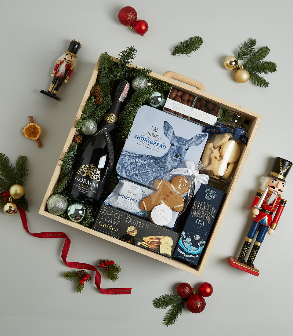 Luxurious Christmas gift basket with Floralba sparkling wine, shortbread, black truffle crisps, Silver Moon tea, chocolate almonds, and a gingerbread cookie in a festive blue and green theme.