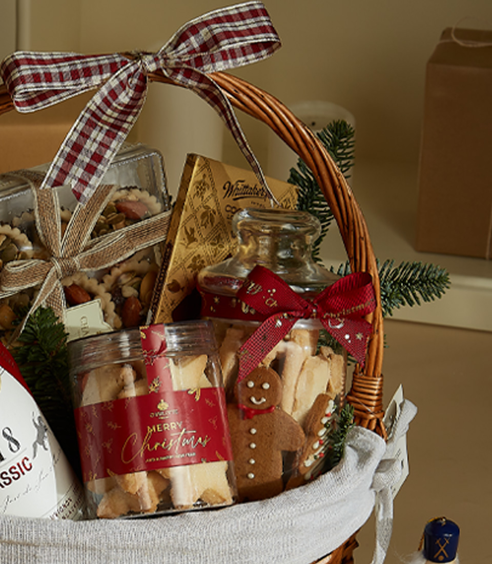Holiday gift basket with 1818 Classic Red Wine, festive shortbread cookies, gourmet chocolates, and snacks in a decorated wicker basket with ribbon and holiday decor.