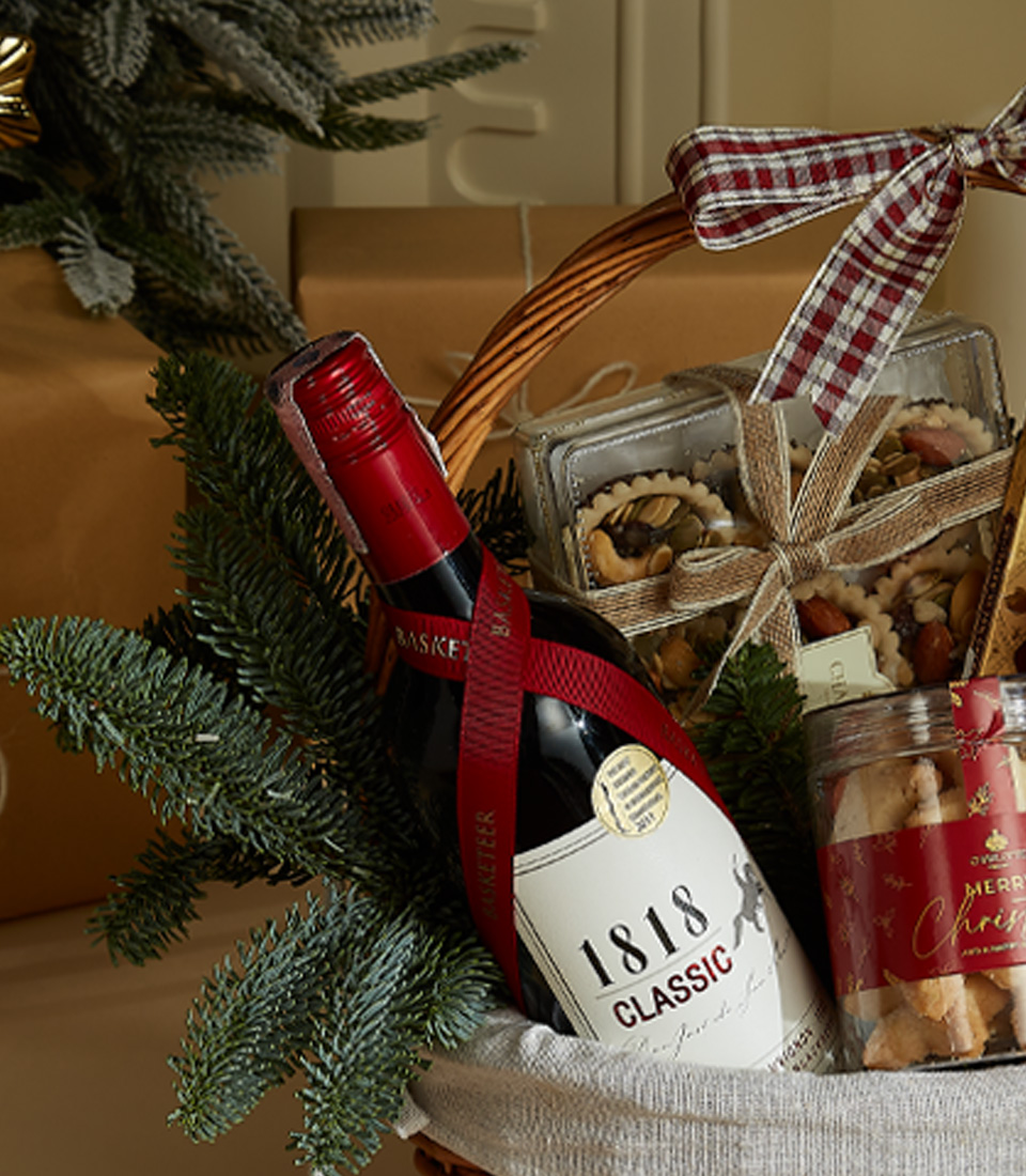 Holiday gift basket with 1818 Classic Red Wine, festive shortbread cookies, gourmet chocolates, and snacks in a decorated wicker basket with ribbon and holiday decor.
