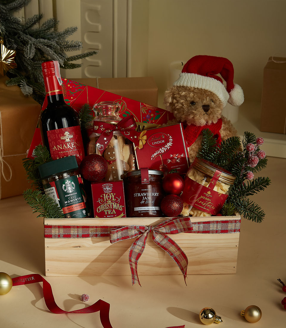 The Christmas Delight Wooden Gift Hamper features a festive array: a bottle of wine, tea, strawberry spread, honey, crackers, and chocolates. This charming selection is adorned with pine branches, baubles, and a Santa-hatted teddy bear in a rustic wooden crate wrapped with a plaid ribbon.