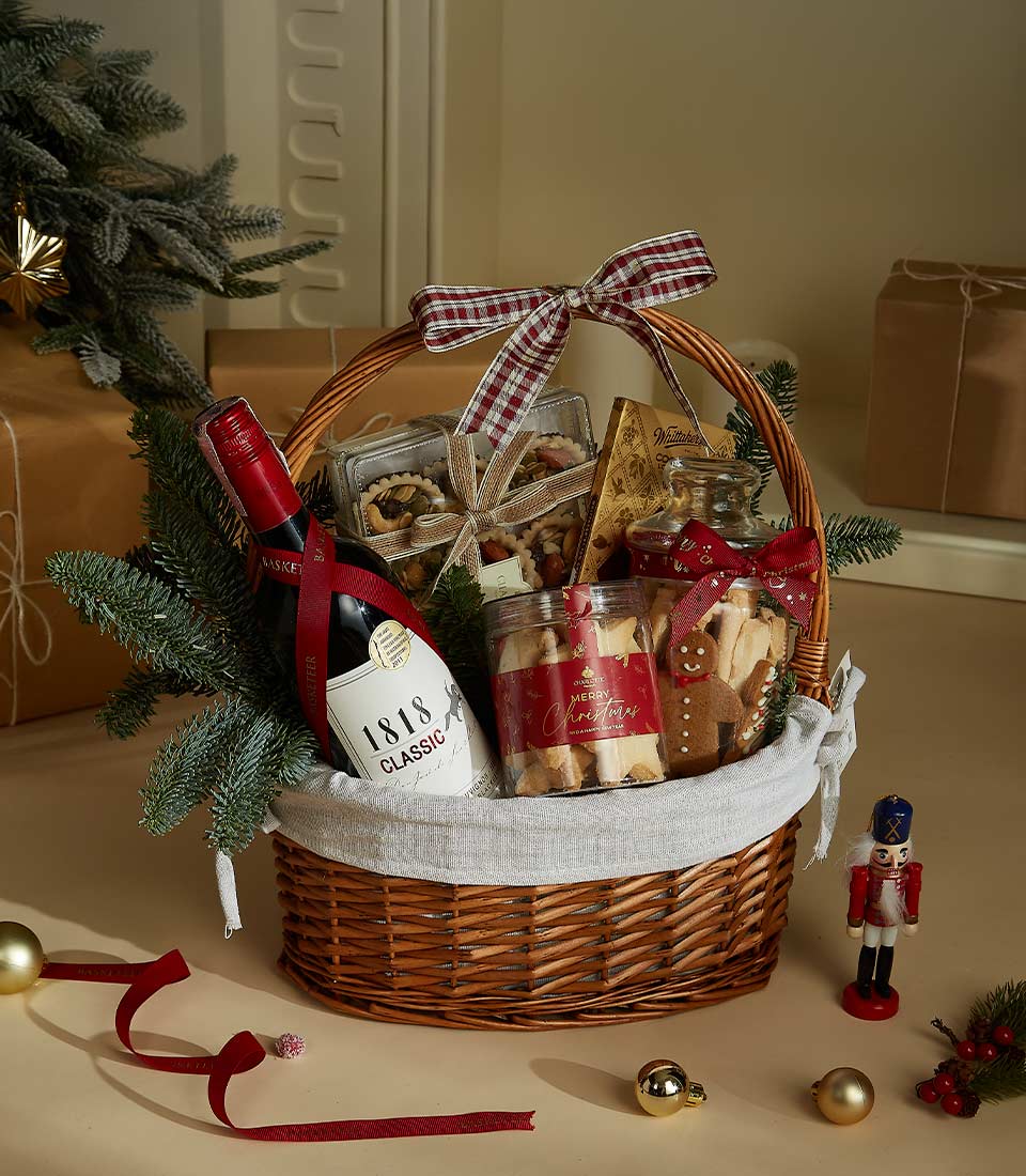 Holiday gift basket with 1818 Classic Red Wine, festive shortbread cookies, gourmet chocolates, and snacks in a decorated wicker basket with ribbon and holiday decor.
