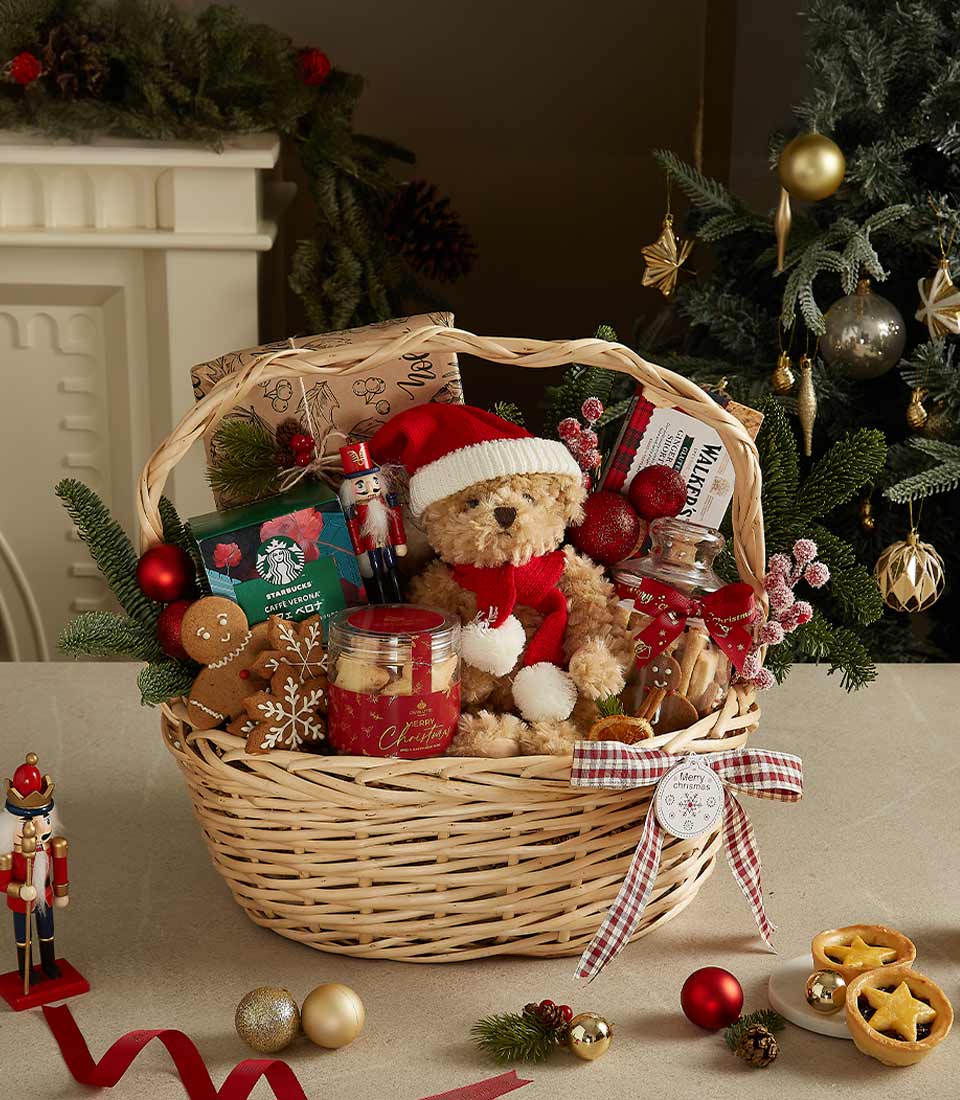 Christmas gift basket with a teddy bear in a Santa hat, tea, festive candle, gingerbread cookies, and holiday decorations.