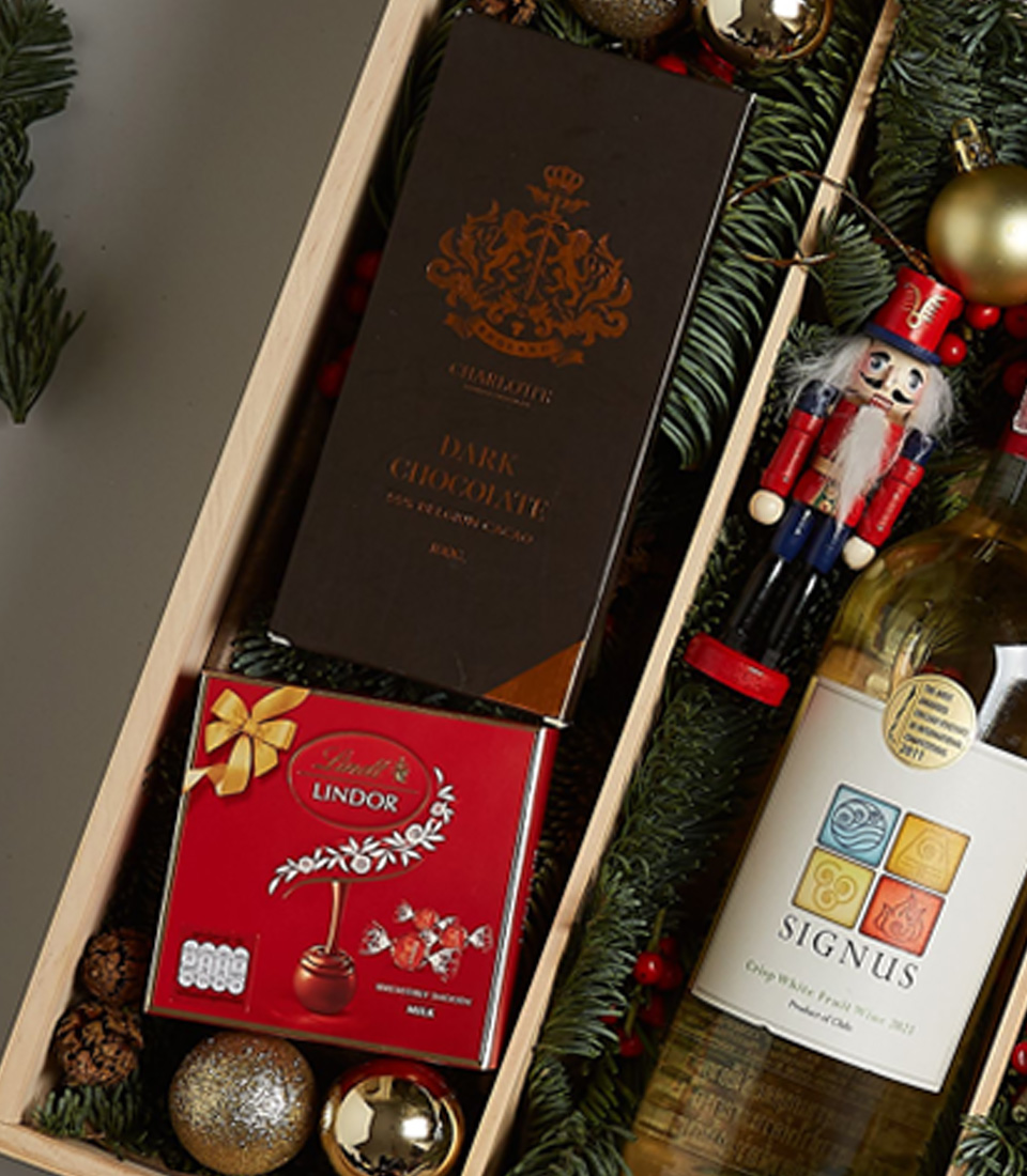Christmas gift box with Signus white wine, dark chocolate, Lindt pralines, cookies, and festive decorations in an elegant arrangement.