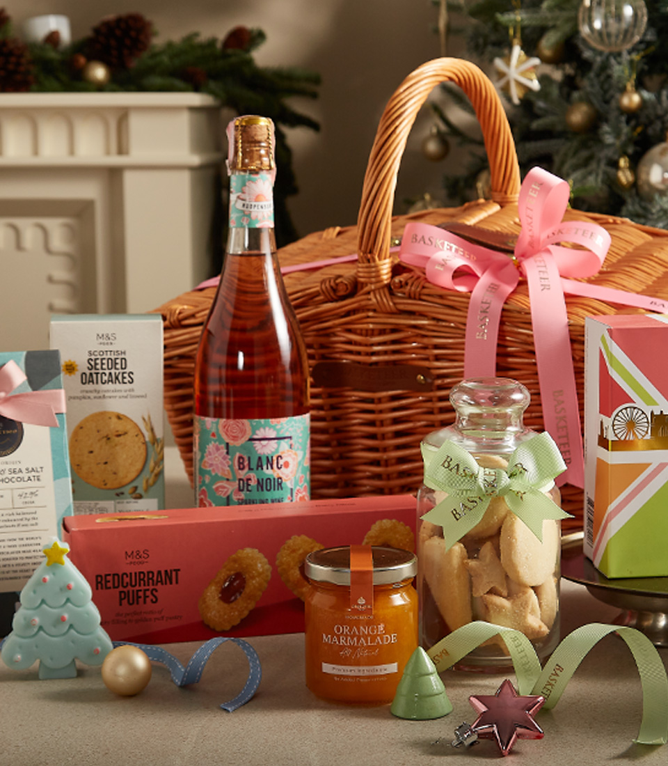 Holiday gift basket with premium tea, sparkling rosé, cookies, and festive decorations in a wicker basket.