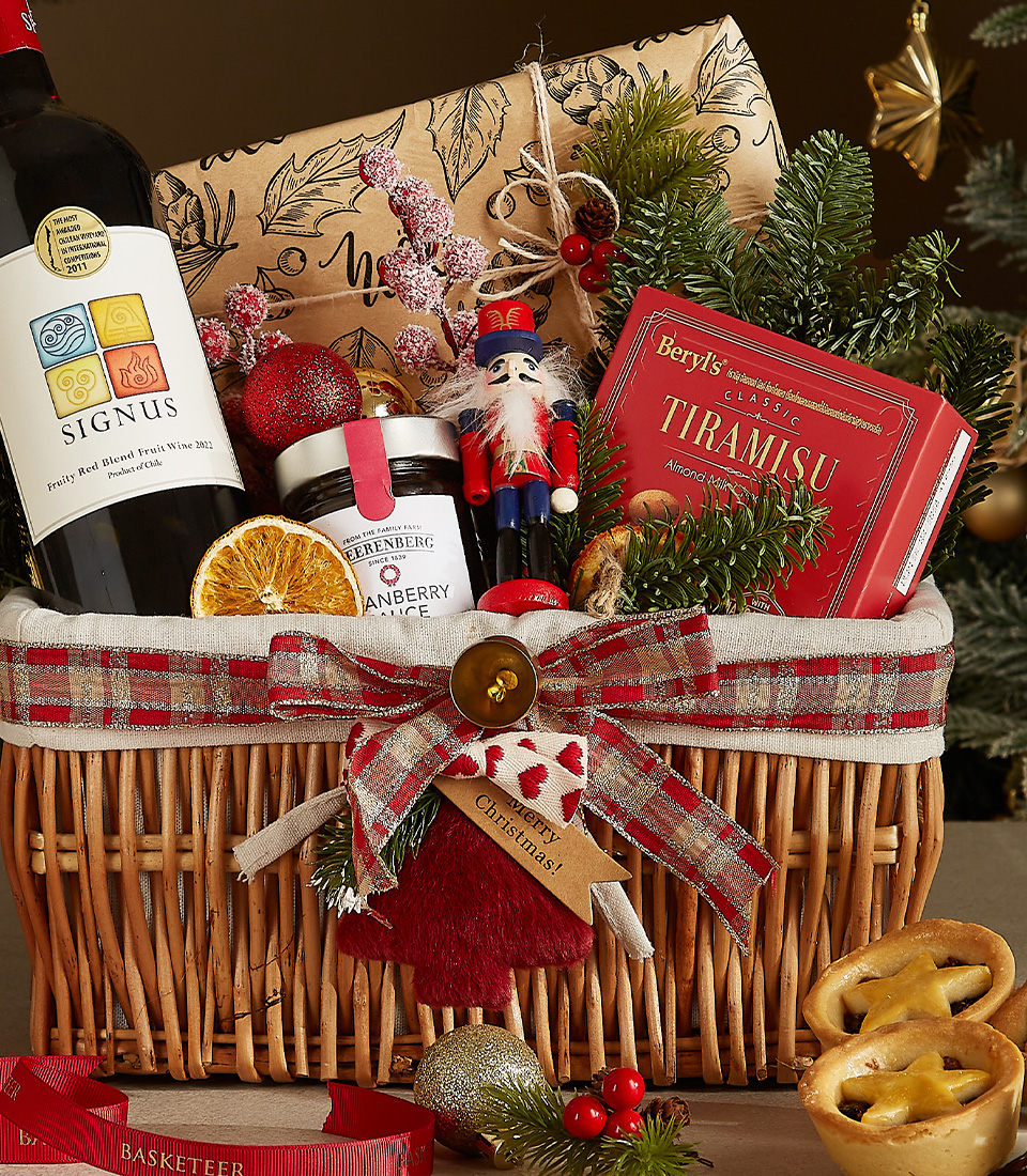 Wicker gift basket containing a bottle of Signus wine, tiramisu chocolates, cranberry sauce, dried orange slices, a nutcracker ornament, and festive greenery.