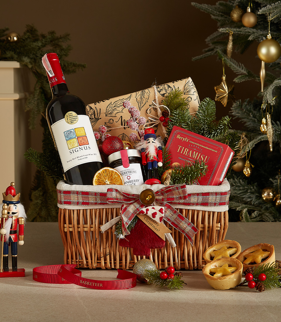 Wicker gift basket containing a bottle of Signus wine, tiramisu chocolates, cranberry sauce, dried orange slices, a nutcracker ornament, and festive greenery.