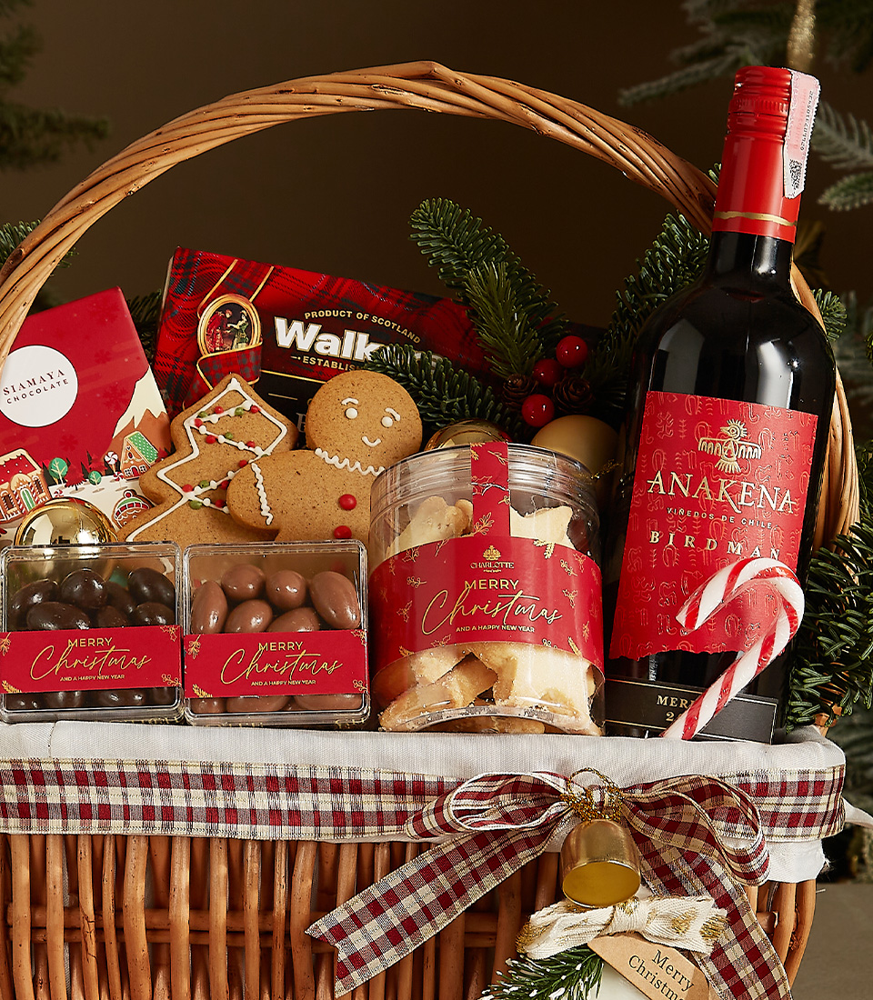 Wicker gift basket containing a bottle of Anakena red wine, shortbread cookies, chocolate almonds, a gingerbread cookie, and festive holiday decorations.