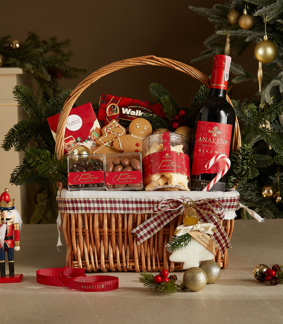 Wicker gift basket containing a bottle of Anakena red wine, shortbread cookies, chocolate almonds, a gingerbread cookie, and festive holiday decorations.