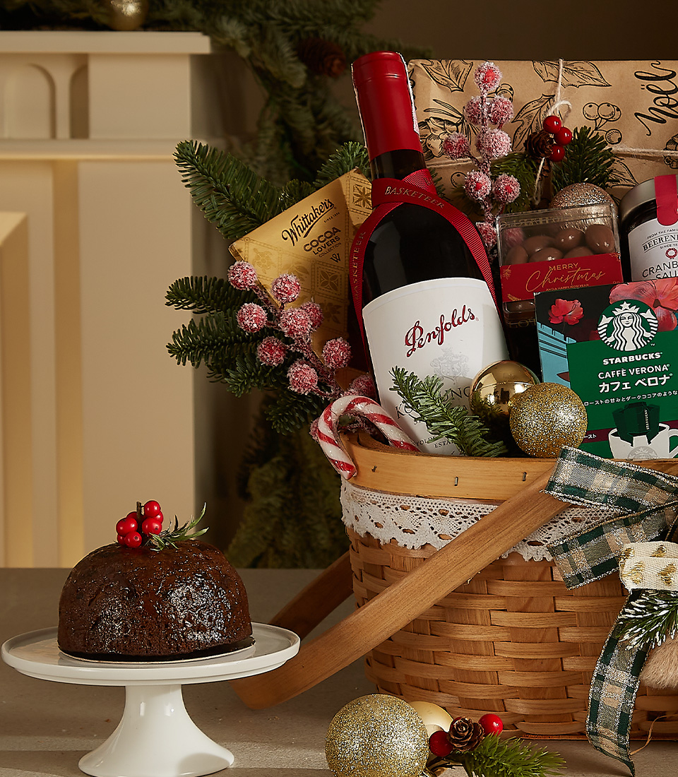 Christmas hamper with red wine, plum pudding, fruit pies, coffee, and holiday-themed treats in a decorated wicker basket.