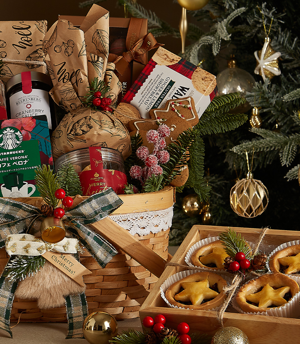 Christmas hamper with red wine, plum pudding, fruit pies, coffee, and holiday-themed treats in a decorated wicker basket.