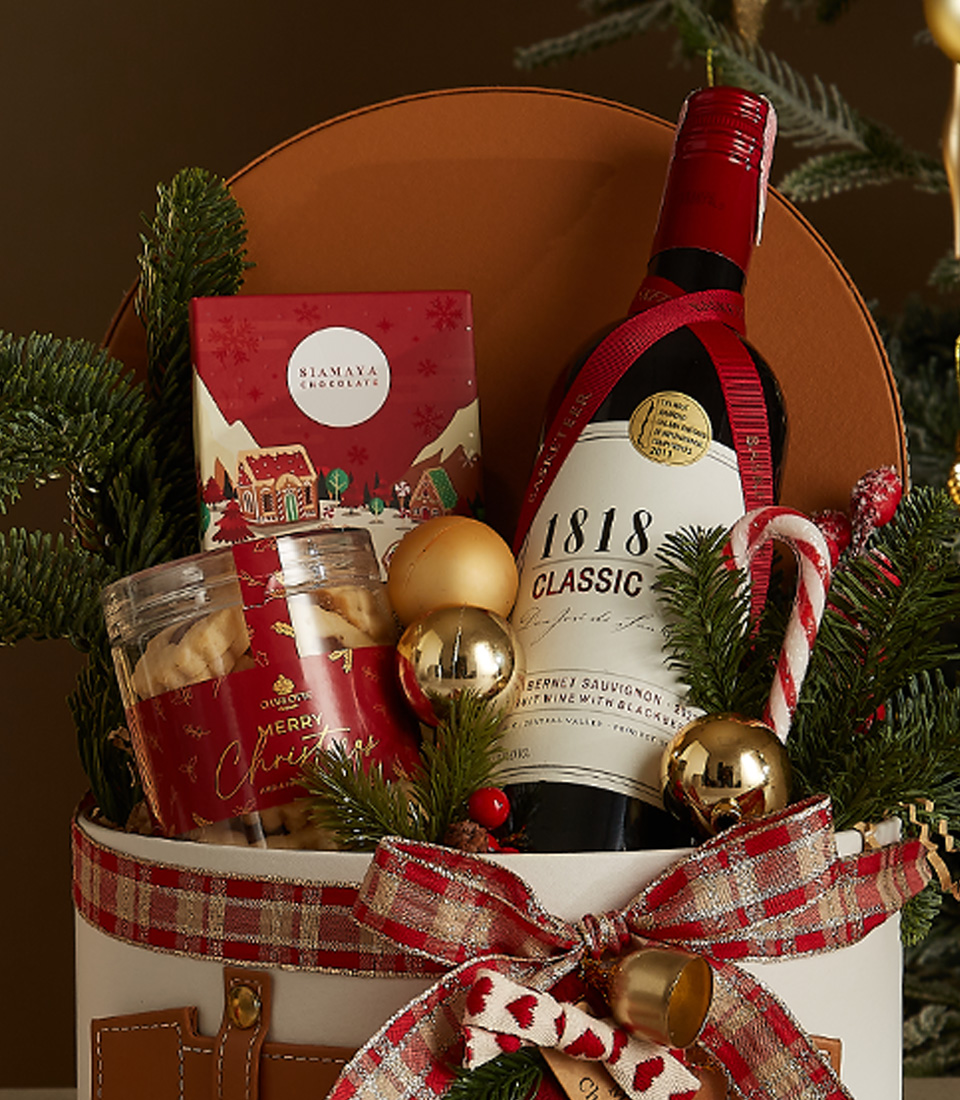 White gift box with leather accents, greenery, and a plaid ribbon containing 1818 Classic Red Wine, butter cookies, chocolates, and a candy cane.