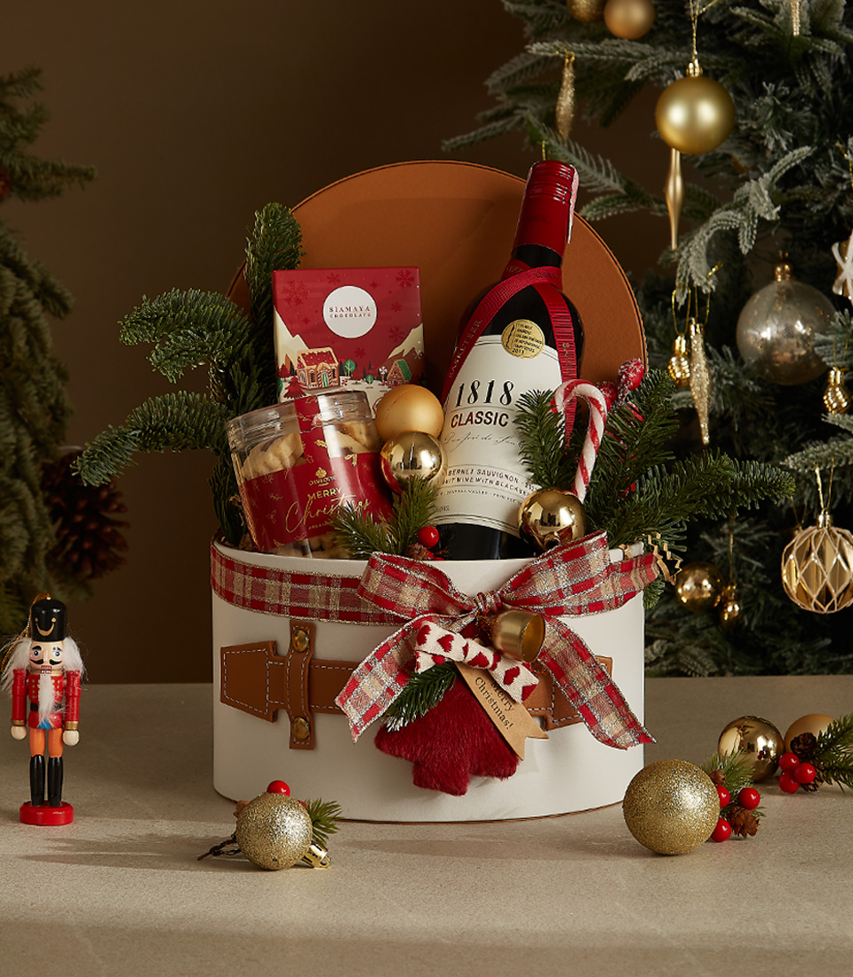 White gift box with leather accents, greenery, and a plaid ribbon containing 1818 Classic Red Wine, butter cookies, chocolates, and a candy cane.