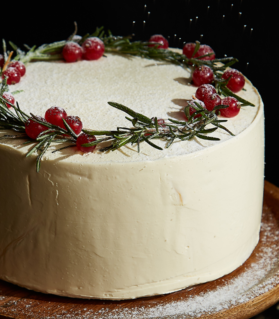 Close-up of Winter Wonderland carrot cake decorated with cream cheese frosting, fresh rosemary sprigs, and cranberries.