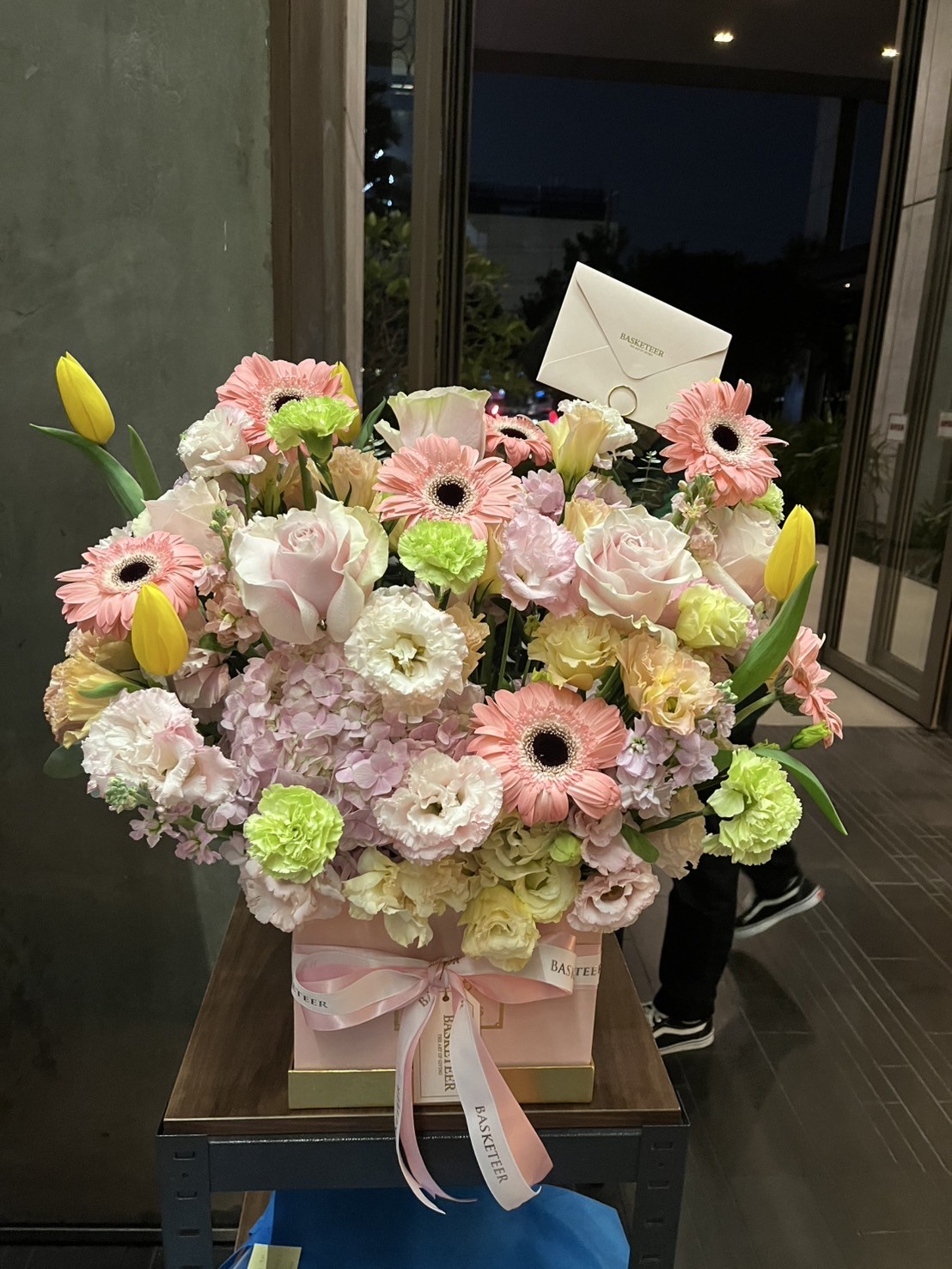 A Radiant Spring Flowers Gift Box featuring pink roses, white and pink gerbera daisies, yellow tulips, and hydrangeas in a box wrapped with a pink ribbon and card, beautifully displayed indoors near a window.