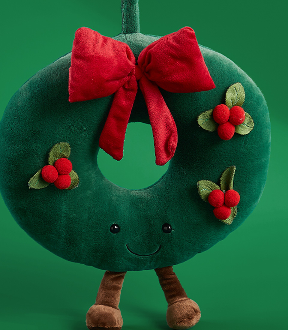 Close-up of a green plush wreath toy with a red bow, holly berry decorations, and smiling face on a green background.