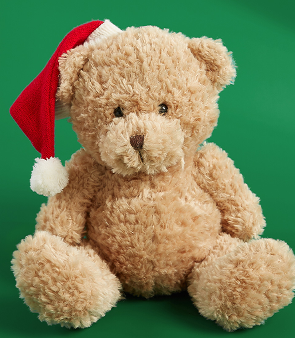 A holiday-themed teddy bear wearing a red Santa hat with a white pom-pom, set against a green background.