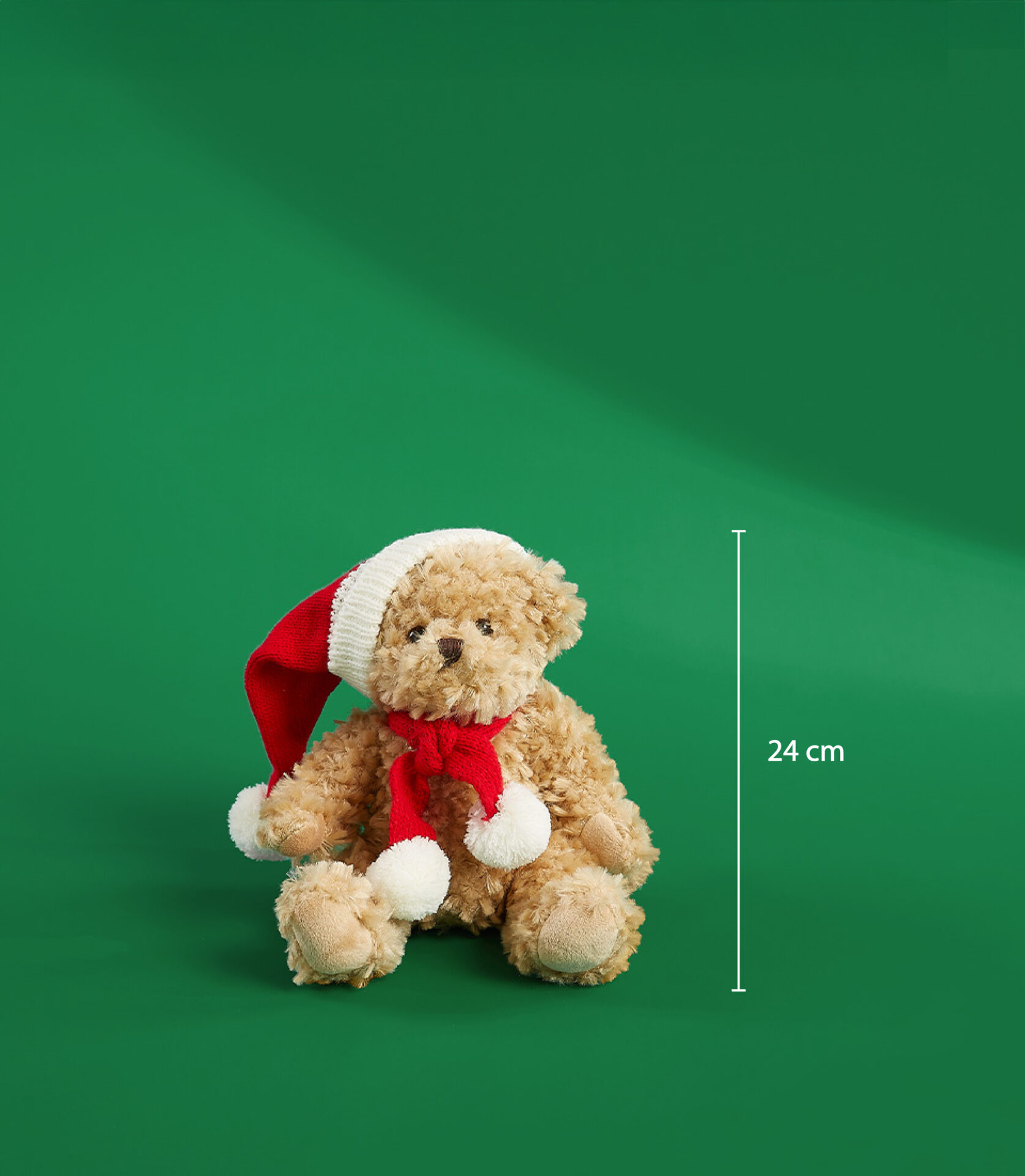 A cute mini teddy bear wearing a red Santa hat and scarf with white pom-poms, sitting on a green background. The bear measures 24 cm.