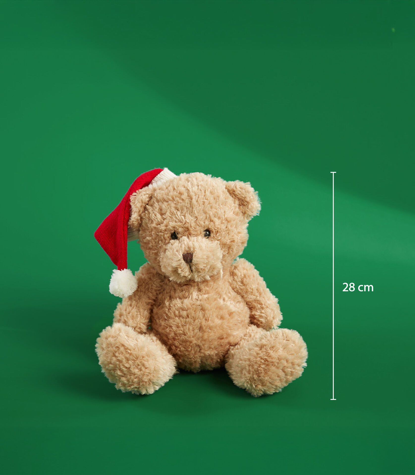 A soft teddy bear wearing a red Santa hat with a white pom-pom, sitting on a green background. The bear measures 28 cm.