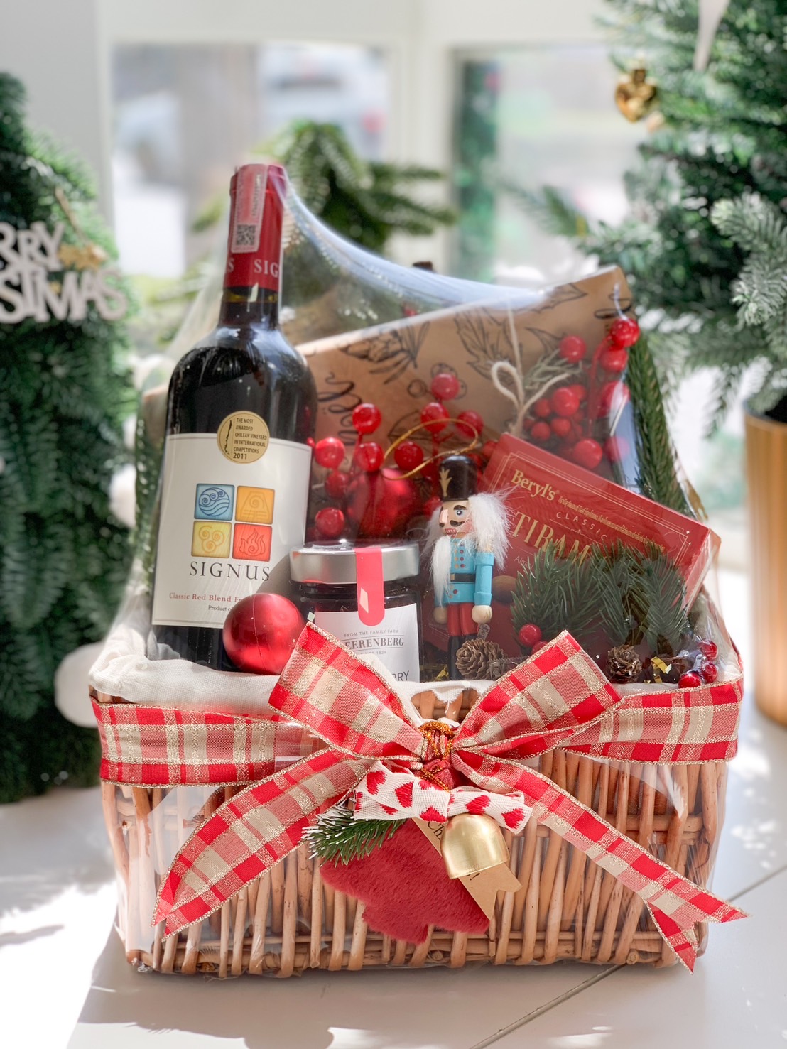 A festive holiday gift basket containing a bottle of wine, chocolates, jam, gourmet crackers, a Nutcracker ornament, and holiday decorations tied with a red plaid ribbon.