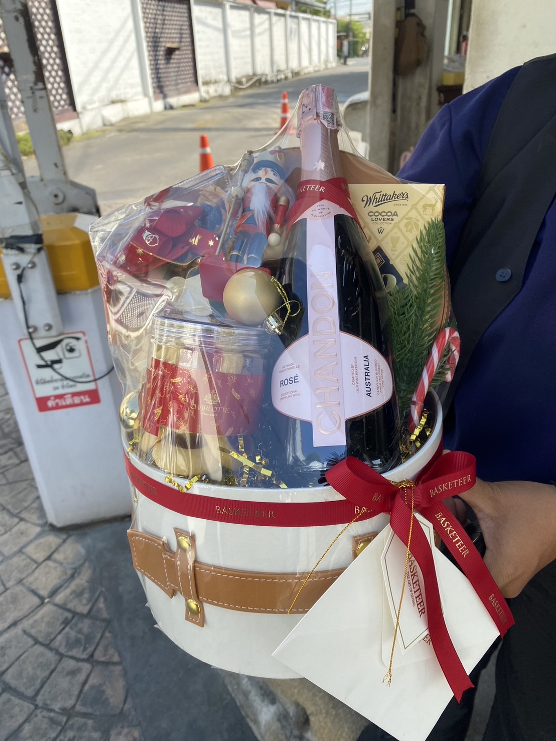 White round gift box with leather strap accents, featuring a bottle of Chandon Rosé, chocolates, festive decorations, and a red Basketeer ribbon.
