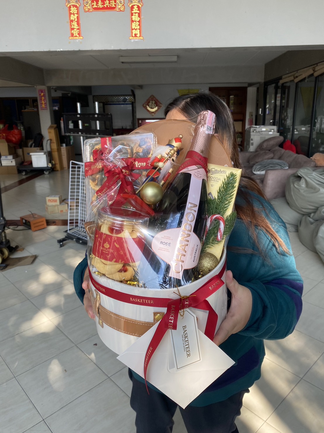 A festive white round gift box with leather accents, containing Chandon Rosé, holiday treats, and seasonal decorations, wrapped with a red Basketeer ribbon.