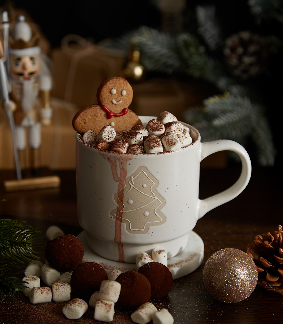 Christmas-themed ceramic mug filled with hot chocolate, topped with marshmallows and a gingerbread cookie, surrounded by chocolate truffles and festive decor.