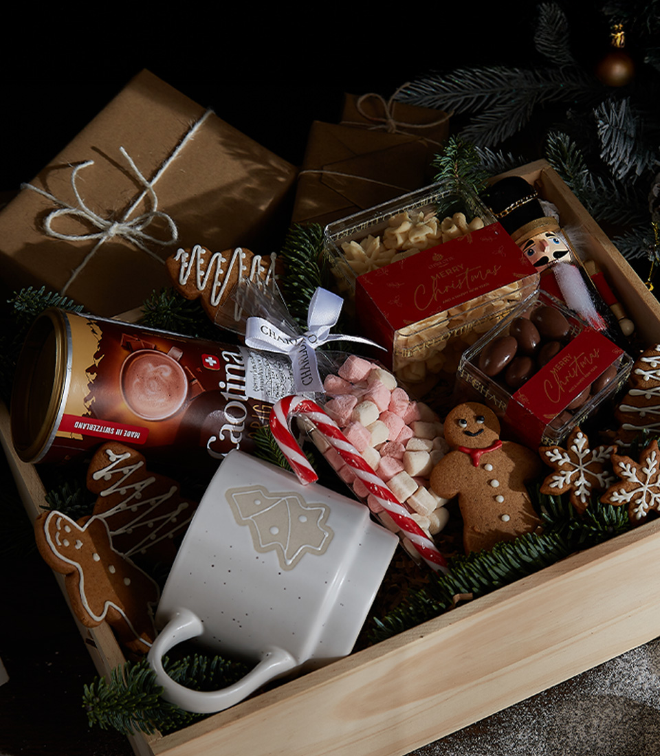 Close-up of a Wooden gift box filled with Caotina hot chocolate, a holiday mug, gingerbread cookies, marshmallows, chocolate-covered nuts, and festive candy canes.