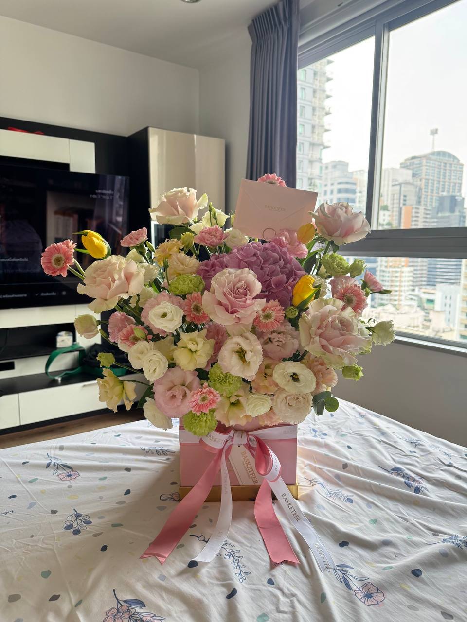 An elegant pink gift box filled with lush spring flowers in pink, white, yellow, and green rests gracefully in a sunlit Bangkok room.