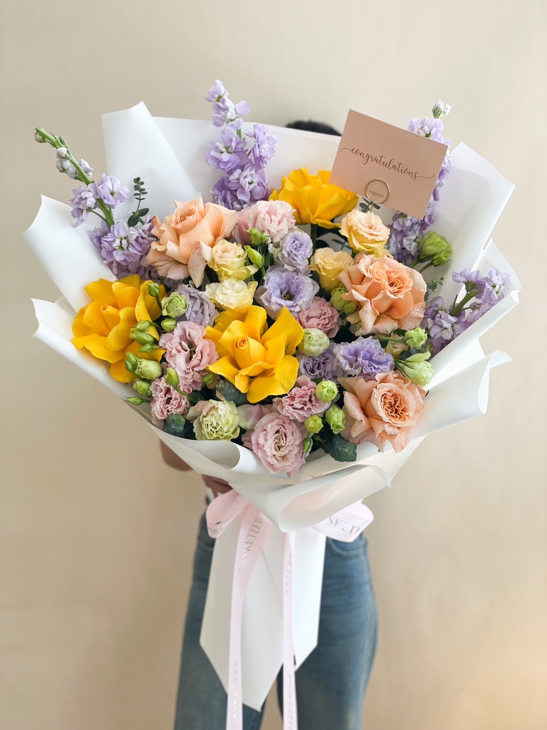 Elegant celebration flower bouquet with yellow, peach, and lavender blooms, wrapped in white paper and finished with a satin ribbon.