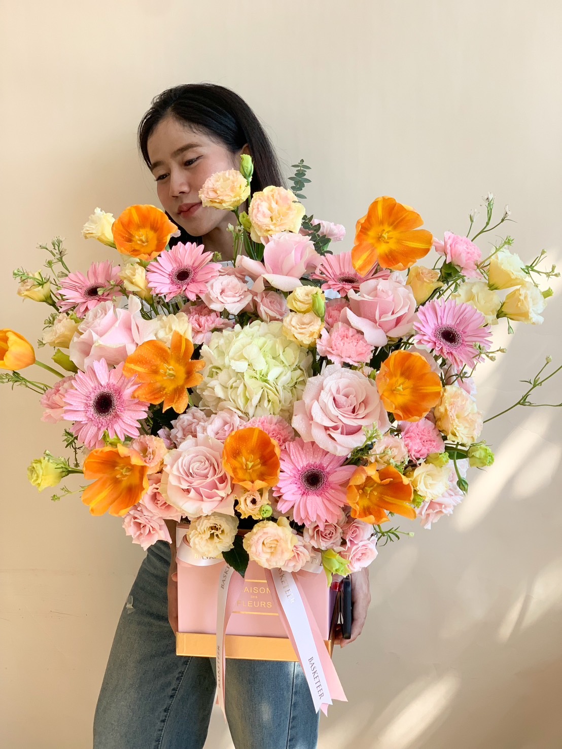 A luxurious floral arrangement in a gift box featuring pink roses, orange poppies, and gerbera daisies, held by a woman.