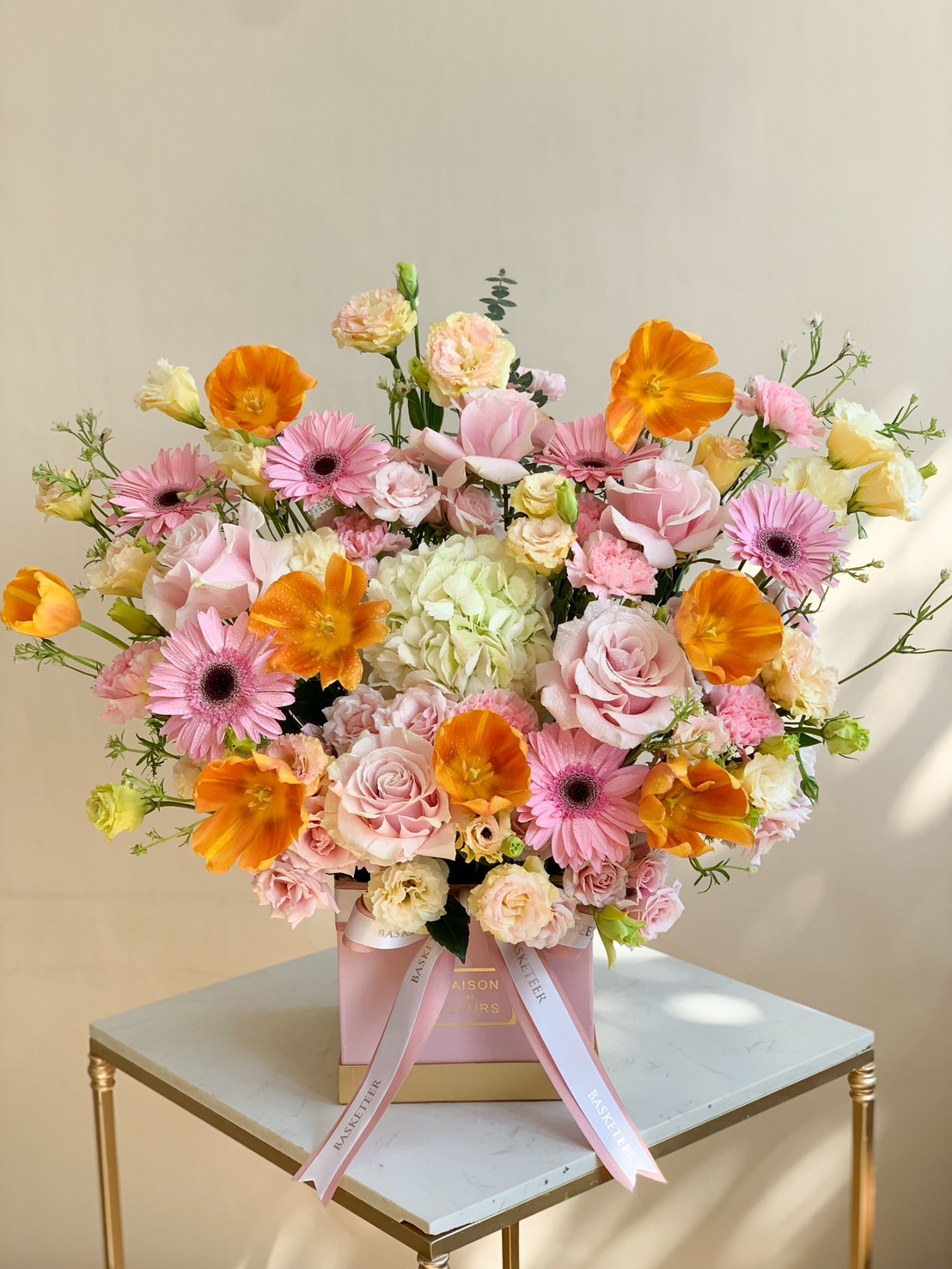 A luxurious floral box arrangement featuring pink roses, orange poppies, gerbera daisies, and hydrangeas, beautifully displayed on a table.