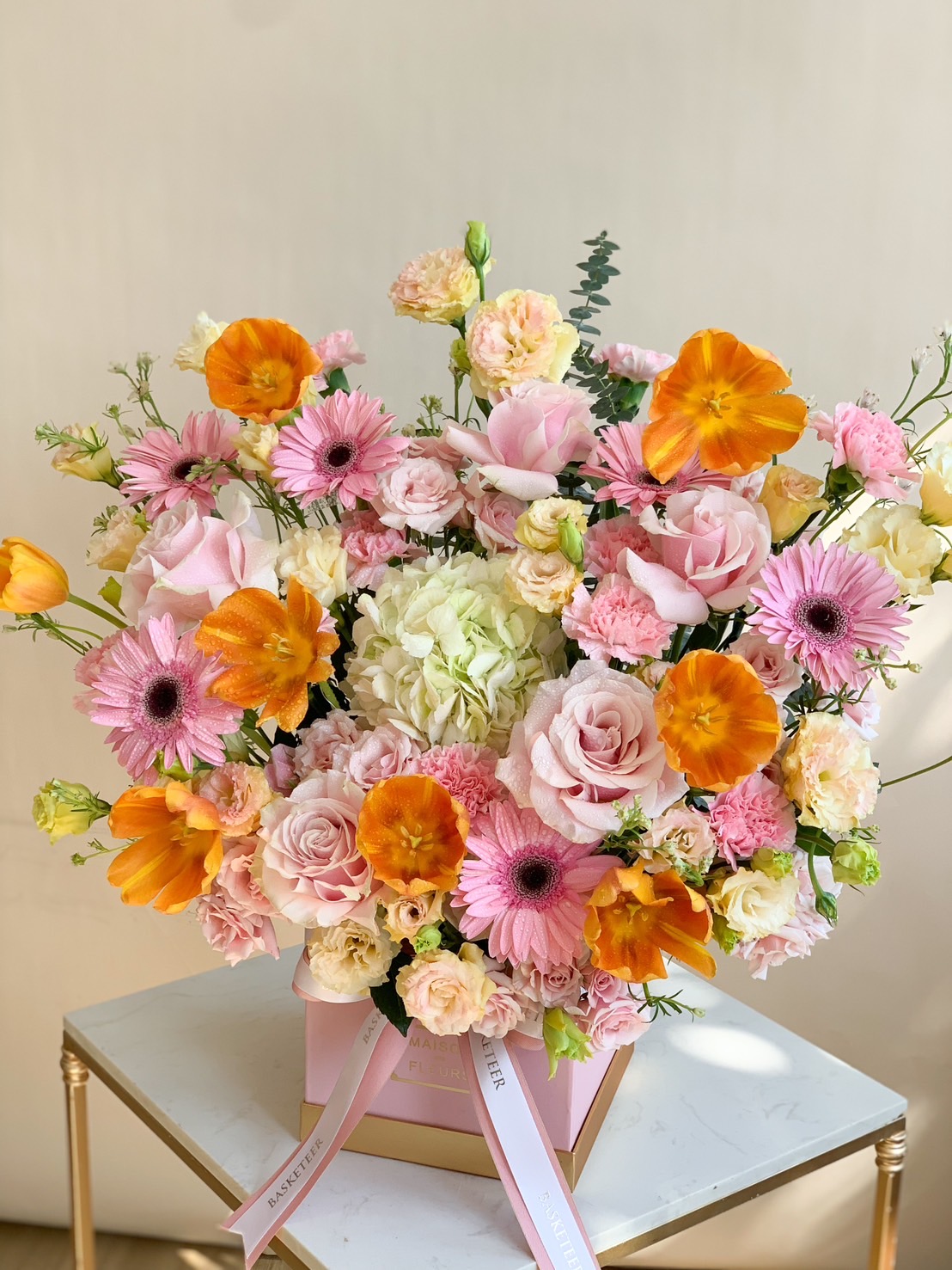 A luxurious floral box arrangement featuring pink roses, orange poppies, gerbera daisies, and hydrangeas, beautifully displayed on a table.