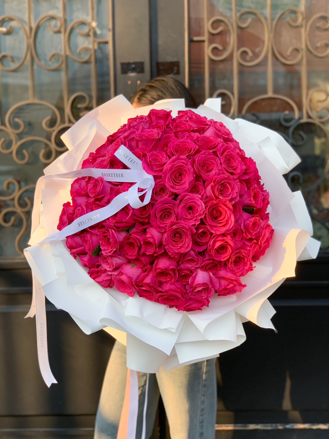 A breathtaking bouquet of 100 hot pink roses, elegantly wrapped with white paper, perfect for special occasions like proposals, anniversaries, and grand romantic surprises.