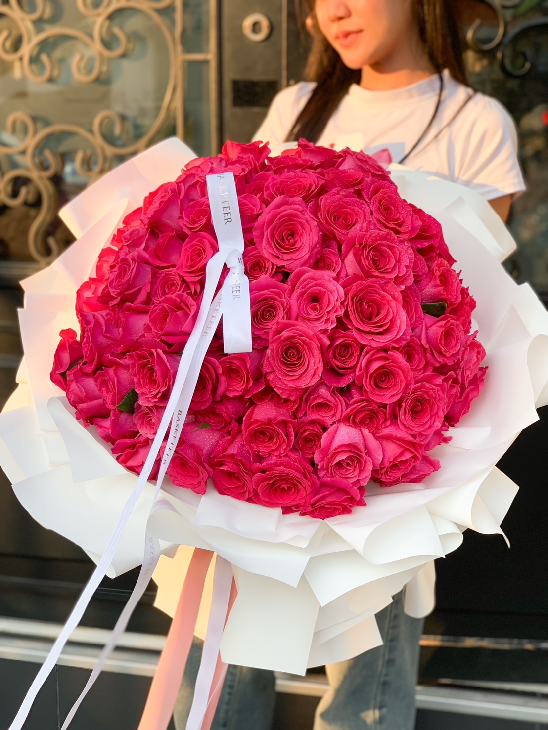 A breathtaking bouquet of 100 hot pink roses, elegantly wrapped with white paper, perfect for special occasions like proposals, anniversaries, and grand romantic surprises.