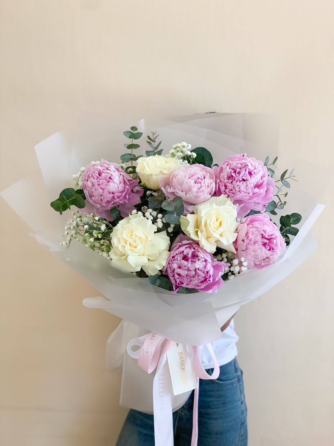 A luxurious hand-tied bouquet featuring delicate pink peonies, ivory roses, eucalyptus leaves, and baby's breath, beautifully wrapped in soft translucent paper with pastel ribbons. A perfect gift for special occasions.